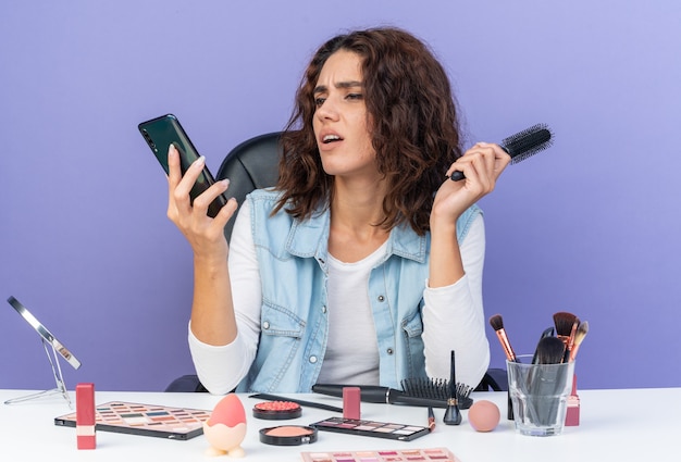 Verärgerte hübsche kaukasische Frau, die am Tisch mit Make-up-Tools sitzt und Kamm hält und auf das Telefon isoliert auf lila Wand mit Kopienraum schaut