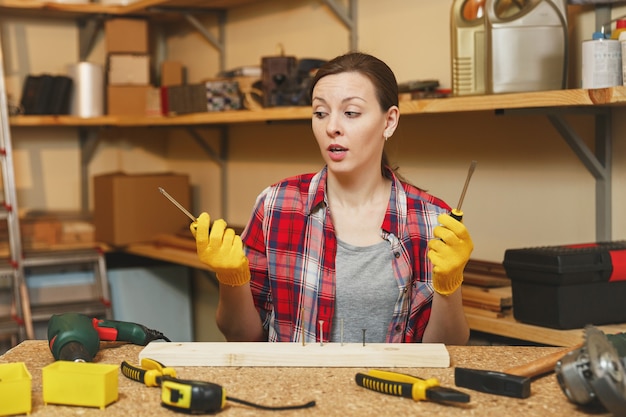 Verärgerte kaukasische junge Frau in kariertem Hemd, grauem T-Shirt, gelben Handschuhen, die sich mit Schraubendreherschraube drehen, in der Tischlerei am Holztisch mit Stück Holz arbeiten, verschiedene Werkzeuge