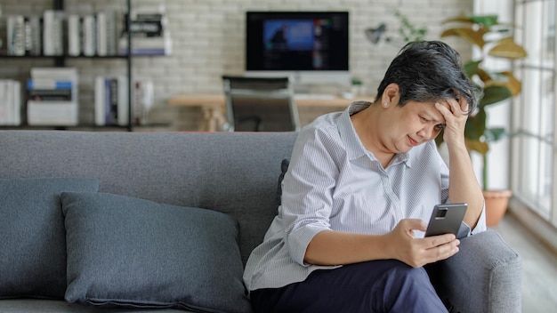 Verärgerte reife Asiatin, die die Stirn runzelt und das Smartphone durchsucht, während sie zu Hause auf dem Sofa im modernen Wohnzimmer sitzt