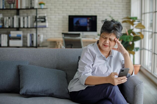 Verärgerte reife Asiatin, die die Stirn runzelt und das Smartphone durchsucht, während sie zu Hause auf dem Sofa im modernen Wohnzimmer sitzt