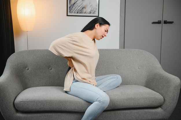 Verärgerte reife Frau, die unter Rückenschmerzen leidet, unglückliche ältere Blondine, die auf einem Sofa im Wohnzimmer sitzt und sich wegen Rückenschmerzen unwohl fühlt