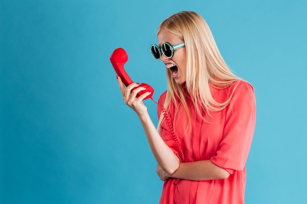 Verärgerte wütende Frau mit Sonnenbrille schreit auf Telefonschlauch isoliert auf einer blauen Wand