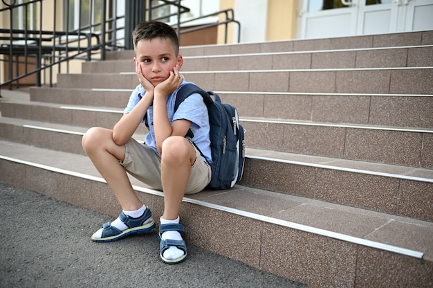 Verärgerter besorgter Junge mit Rucksack, der auf der Treppe von der Schule sitzt. Mobbing, Einsamkeit, Lernschwierigkeiten. Zurück zum Schulkonzept