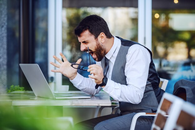 Verärgerter eleganter Geschäftsmann im Anzug, der Laptop betrachtet und schreit, während er im Café sitzt.