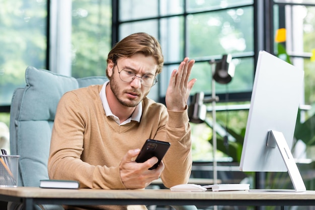 Verärgerter junger männlicher freiberuflicher Programmierer, der im Büro am Tisch sitzt und das Telefon in seinem hält