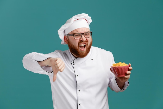 Verärgerter junger männlicher Koch mit Brille, Uniform und Mütze, der in die Kamera blickt und eine Schüssel Makkaroni zeigt, die den Daumen nach unten zeigt, isoliert auf blauem Hintergrund