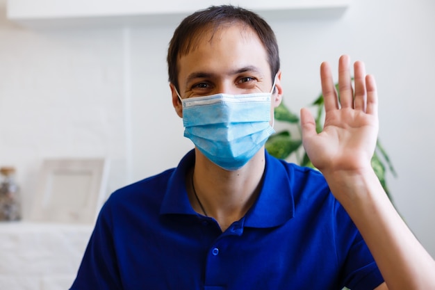 Verärgerter junger Mann in grauem Pullover, Schal mit steriler Gesichtsmaske, der Videoanrufe mit dem Handy tätigt, Hand auf grauem Hintergrund ausbreitend. Gesundheit, Behandlung kranker Krankheiten, Konzept der kalten Jahreszeit