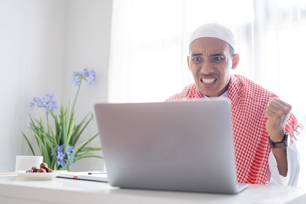 Verärgerter muslimischer Mann beim Betrachten seines Laptopbildschirms, der auf seinem Schreibtisch sitzt