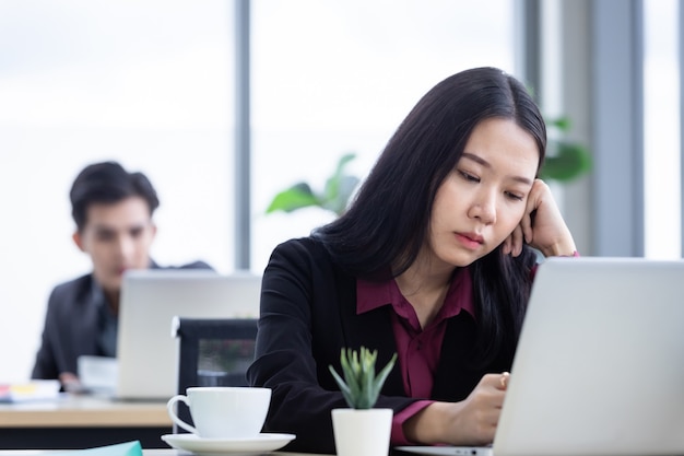 Verärgerter nachdenklicher Stress der jungen asiatischen Geschäftsfrau am Arbeitsplatz, der mit Laptop-Computern von Büromitarbeitern arbeitet, ist nicht glücklich mit Arbeitskollegen im Büro