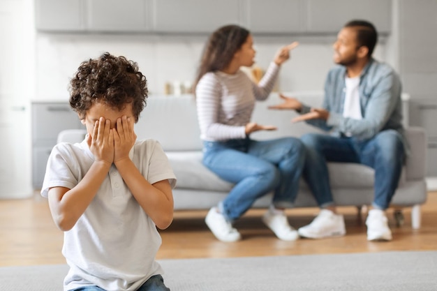 Verärgerter schwarzer Preteen-Junge, der sein Gesicht mit den Händen bedeckt, während seine Eltern streiten