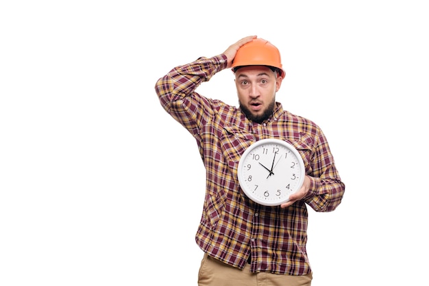 Verärgerter und geschockter Baumeisterarbeiter in der Schutzkonstruktion orange Helm, der in der Hand einen großen Wecker hält, der auf weißem Hintergrund lokalisiert wird. Zeit zu Arbeiten. Bauzeit.
