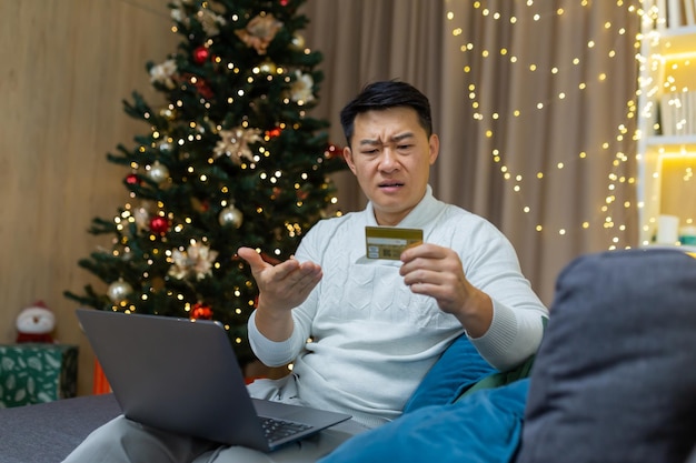 Verärgerter weihnachtsmann mit laptop, der versucht, online im online-shop einzukaufen asiatischer mann hält