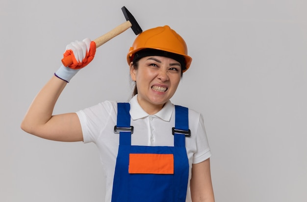 Verärgertes junges asiatisches Baumeistermädchen mit orangefarbenem Schutzhelm und Sicherheitshandschuhen, die Hammer halten