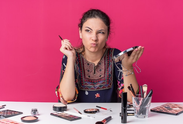 Foto verärgertes junges brünettes mädchen, das am tisch mit make-up-tools, make-up-pinsel und spiegel sitzt und auf die seite isoliert auf rosa wand mit kopierraum schaut