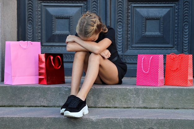 Verärgertes junges Mädchen sitzt mit bunten Taschen auf den Stufen des Ladens