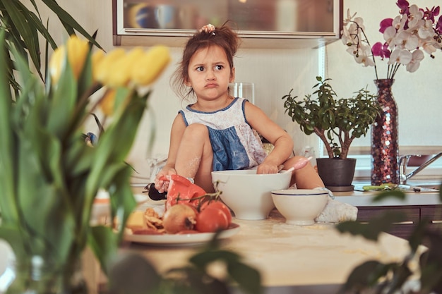 Verärgertes kleines süßes Mädchen lernt, eine Mahlzeit in der Küche zu kochen, während es auf einer Arbeitsplatte sitzt.