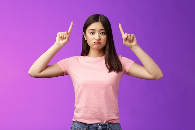 Verärgertes, launisches, trauriges asiatisches Mädchen sieht eifersüchtig aus, schmollend beunruhigt, zeigt nach oben, zeigt Zeigefinger Top-Werbung bedauert verpasste Chance, steht enttäuscht lila Hintergrund unzufrieden