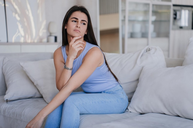 Verärgertes spanisches Mädchen in lila Bluse und blauen Jeans sitzt zu Hause auf dem Sofa und blickt zur Seite