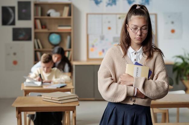 Verärgertes und einsames Schulmädchen mit Heften, die vor der Kamera stehen