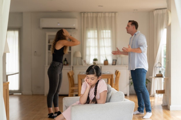 Verärgertes und unglückliches junges Mädchen, das auf dem Sofa sitzt und durch den Streit ihrer Eltern im Wohnzimmer mitten in der Anspannung gefangen ist. Ungesunder häuslicher Lebensstil und traumatische Kindheit entwickeln sich zu DepressionenSynchronos