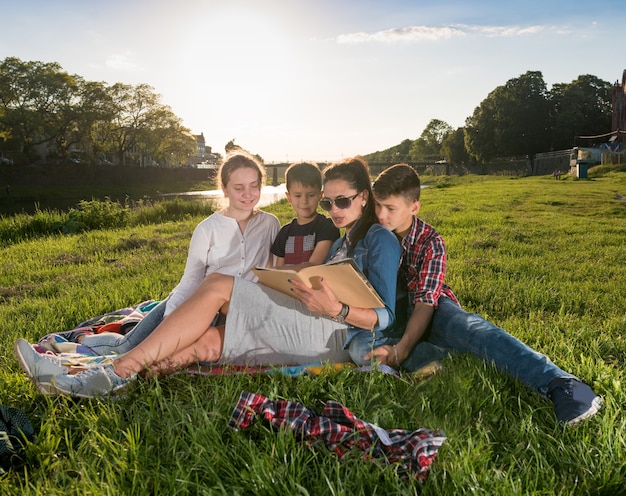 Verano, infancia, concepto de ocio. Madre con niños sentados en el césped y leyendo un libro en el parque. Día soleado de verano