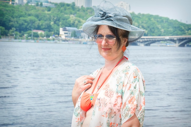 En verano, una mujer imponente con sombrero y gafas se encuentra cerca del río.