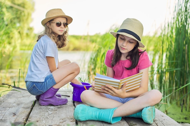 Verano, vacaciones, naturaleza, estilo de vida, ocio infantil. Niños, dos niñas sentadas en un muelle de madera en un lago de caña, hablando y leyendo un cuaderno con interés