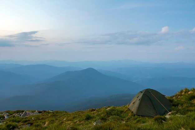 Verão acampar nas montanhas ao amanhecer. Barraca do turista na colina gramada redonda na cordilheira azul enevoada distante sob céu rosa antes do nascer ou pôr do sol. Turismo, caminhadas e beleza do conceito de natureza.