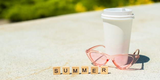 Verão em cubos de madeira, óculos de sol rosa e xícara de café branco sobre fundo branco
