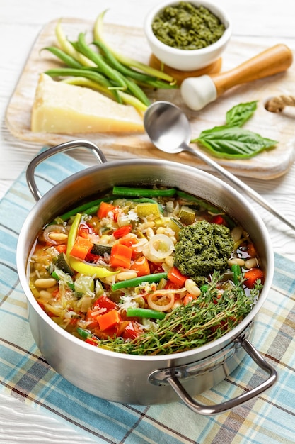 Verão vegetariano Soupe au Pistou sopa de feijão francês com molho pesto e ervas em uma panela de metal em uma mesa de madeira branca com vista vertical de ingredientes