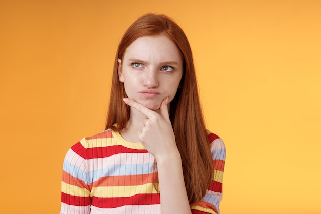 Verblüfft zögernd ernsthaft aussehende Ingwer Mädchen blaue Augen weihing Entscheidungen schmollen Touch Kinn Blick obere linke Ecke nachdenklich denken verwirrte Lösung, Nachdenken Entscheidung, orange Hintergrund.