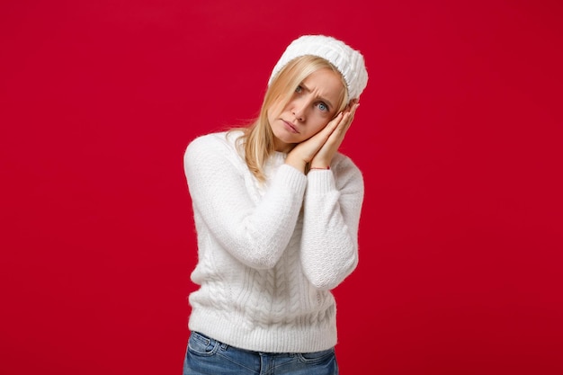 Foto verblüffte junge frau im weißen pullover, hut isoliert auf rotem hintergrund, studioporträt. gesunder modelebensstil, konzept der kalten jahreszeit. mock-up-kopienbereich. versuchen sie, mit gefalteten händen unter der wange zu schlafen.