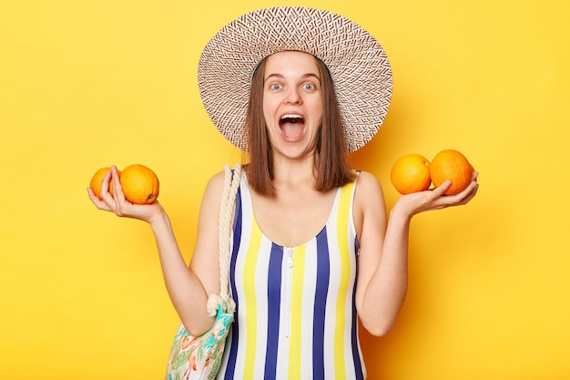 Verblüffte, überraschte, fröhliche Frau mit gestreiftem Badeanzug und Strohhut, isolierter gelber Hintergrund, hält frische süße Orangen in der Hand und genießt Resort und tropische Früchte