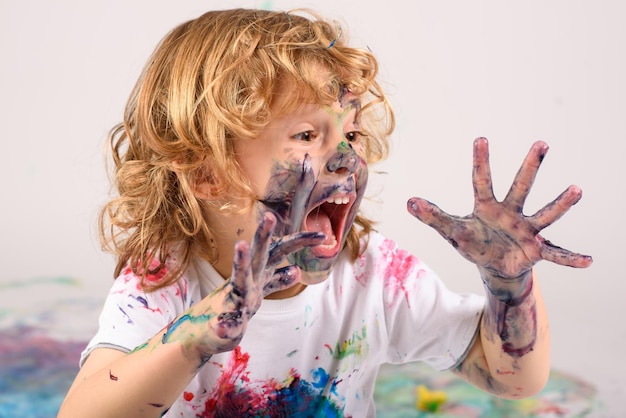 Foto verblüffter, schlampiger junge, bedeckt mit bunten farben, sitzt auf unordentlichem boden mit geöffnetem mund auf weißem hintergrund im hellen studio