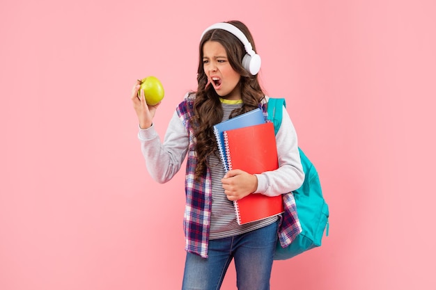 Verblüfftes Teenie-Mädchen mit Rucksack und Arbeitsbuch mit Kopfhörern schauen sich das Apple-Mittagessen an