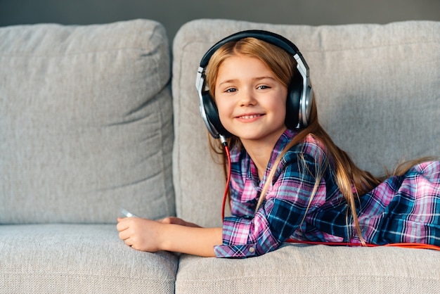 Verbringen Sie tolle Zeit zu Hause. Schönes kleines Mädchen mit Kopfhörern, das mit einem Lächeln in die Kamera schaut, während es zu Hause auf der Couch liegt