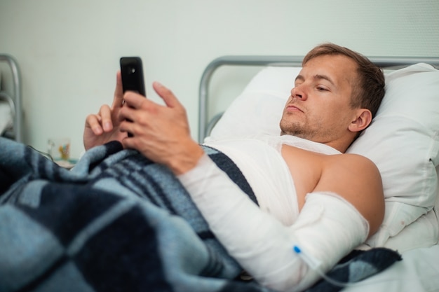 Verbundener Patient, der auf dem Bett im Krankenhaus mit einem Smartphone in seinen Händen liegt