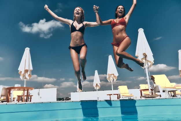 Verdadera alegría. Longitud total de dos atractivas mujeres jóvenes en bikini tomados de la mano y saltando juntos en la piscina