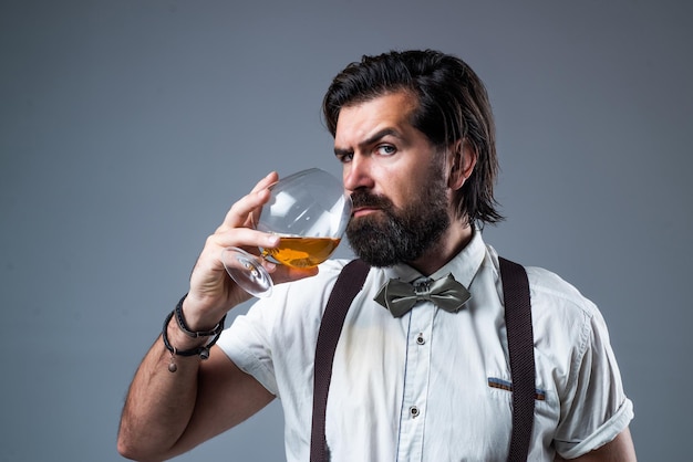 Verdadero caballero con alcohol brandy o coñac beber whisky escocés hombre barbudo en tirantes bebiendo ron vidrio elegante hombre de negocios usar pajarita para evento formal sommelier