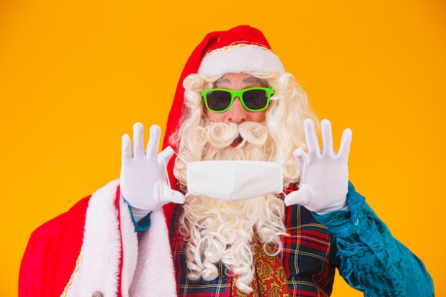 Verdadero Santa Claus sobre fondo amarillo sosteniendo en las manos una máscara protectora contra covid19. Navidad con distancia social. COVID-19