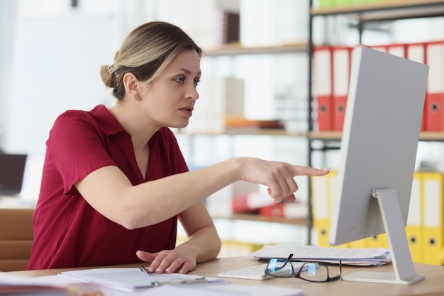 Foto verdächtige frau zeigt mit dem finger auf den bildschirm des computermonitors, der am tisch in der büroblondine sitzt