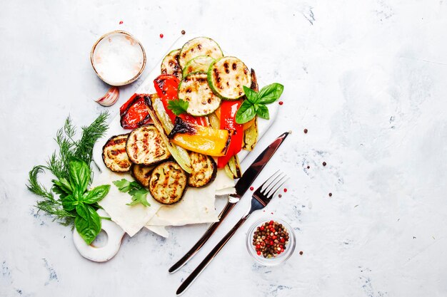 Verduras de colores a la parrilla, berenjenas, pimiento de calabacín con especias y albahaca verde en la tabla de servir en la vista superior de fondo blanco