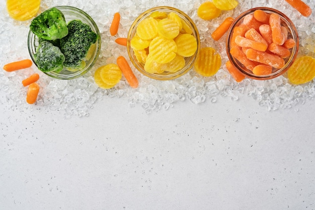 Verduras congeladas como brócoli verde, zanahoria amarilla y baby en tazones de vidrio sobre hielo y mesa gris de hormigón con espacio de copia.