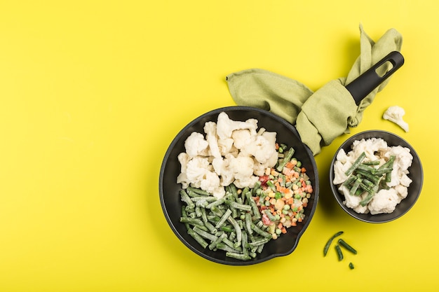 Verduras congeladas: judías verdes, coliflor y una mezcla de verduras en una sartén negra con una servilleta y en un plato sobre un fondo amarillo brillante.