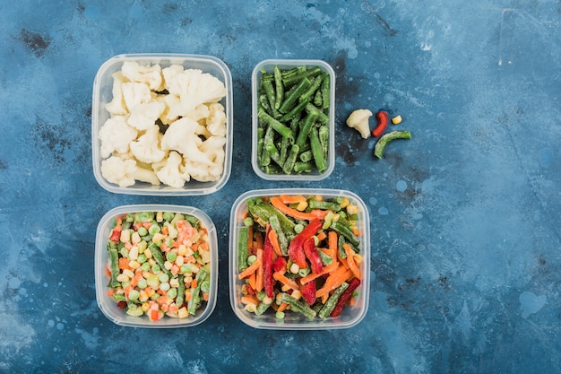 Verduras congeladas: una mezcla de verduras, judías verdes y coliflor en diferentes recipientes de plástico para congelar sobre un fondo azul.