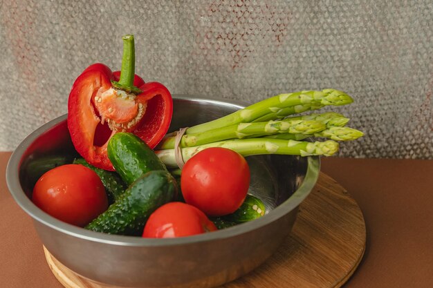 Las verduras se encuentran en un tazón de metal, tomates, espárragos, pepinos, pimientos rojos sobre una tabla de madera y fondo marrón, fondo gris, lugar para el texto