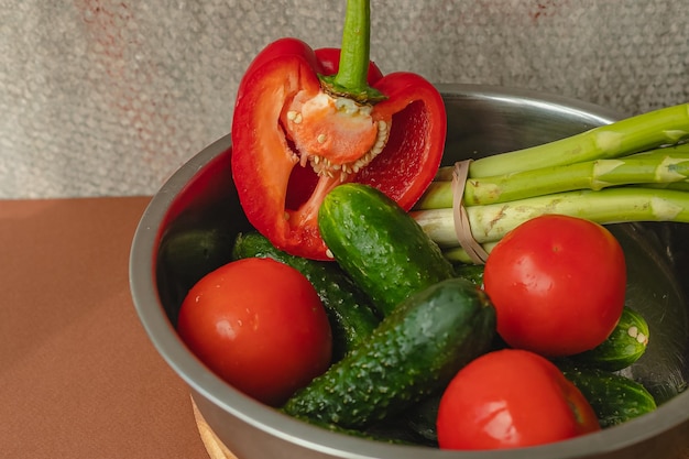 Las verduras se encuentran en un tazón de metal, tomates, espárragos, pepinos, pimientos rojos sobre una tabla de madera y fondo marrón, fondo gris, lugar para el texto