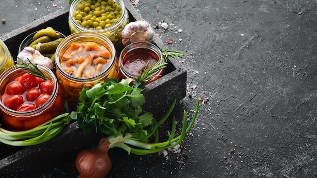 Verduras en escabeche y champiñones en frascos de vidrio en caja de madera sobre fondo de piedra negra Vista superior