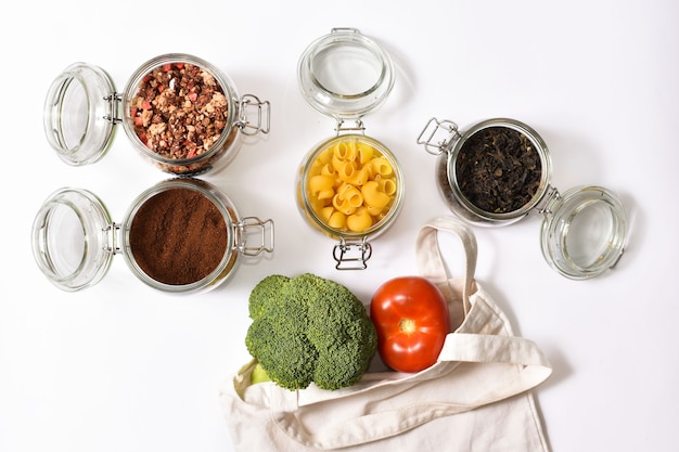 Verduras frescas en una bolsa de asas beige ecológica en la pared blanca. Frascos de vidrio con té verde, granola, café, pasta. Concepto de desperdicio cero, rotundamente.