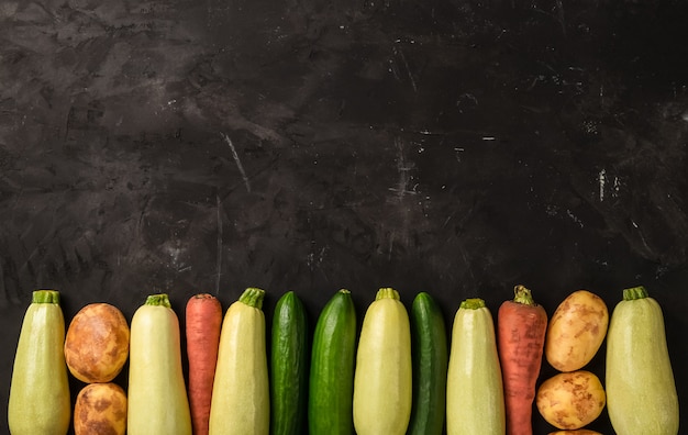 Foto verduras frescas crudas sobre el fondo negro en línea en la vista superior. calabacín, zanahoria, pepino y patata